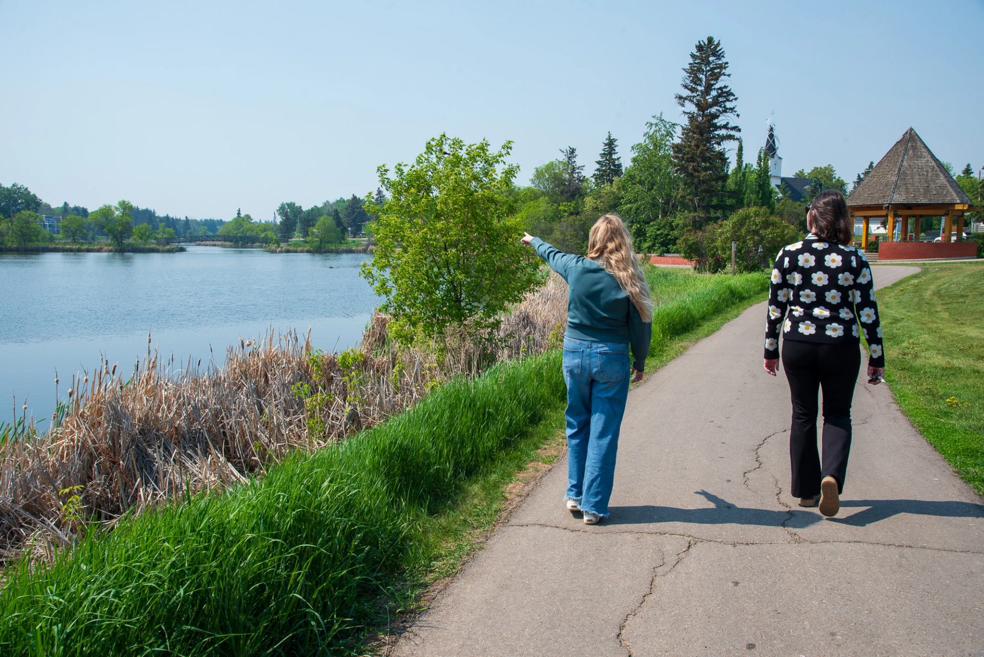 2 friends walking along a paved path by Mirror Lake in Camrose, one of the friends is pointing at a bird.