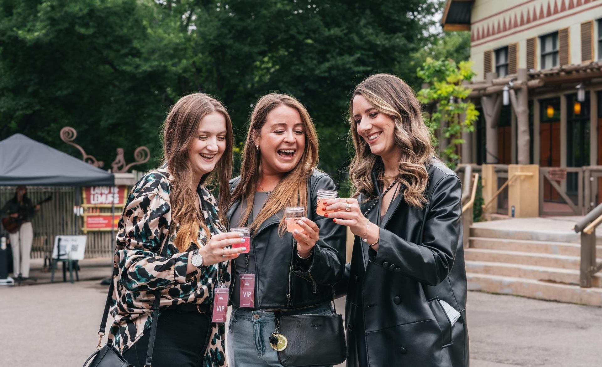 Three adults wearing VIP passes hold small drinks together outside near trees and buildings.