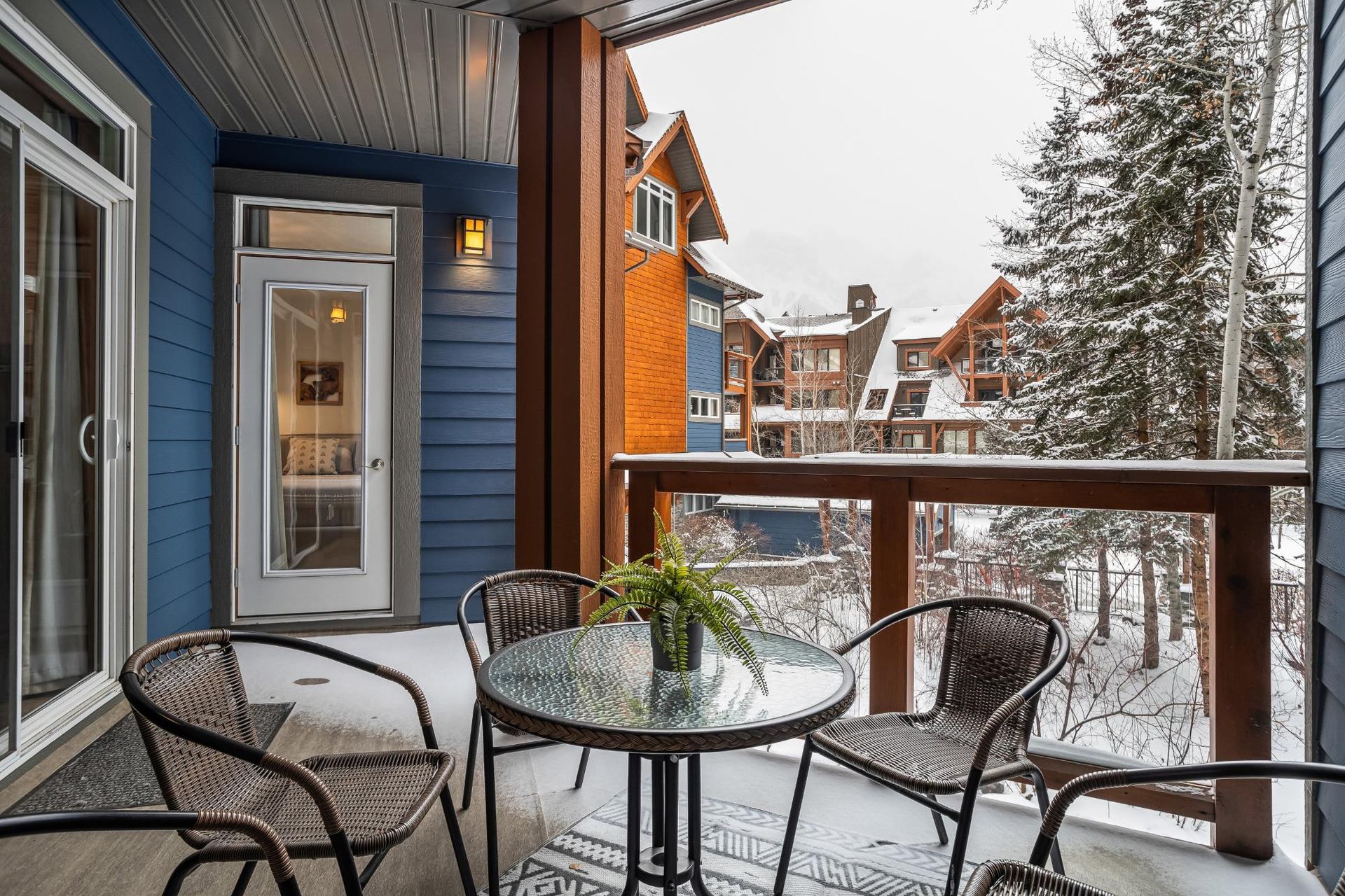 Covered balcony with wicker chairs, glass table, and snowy lodge buildings beyond.