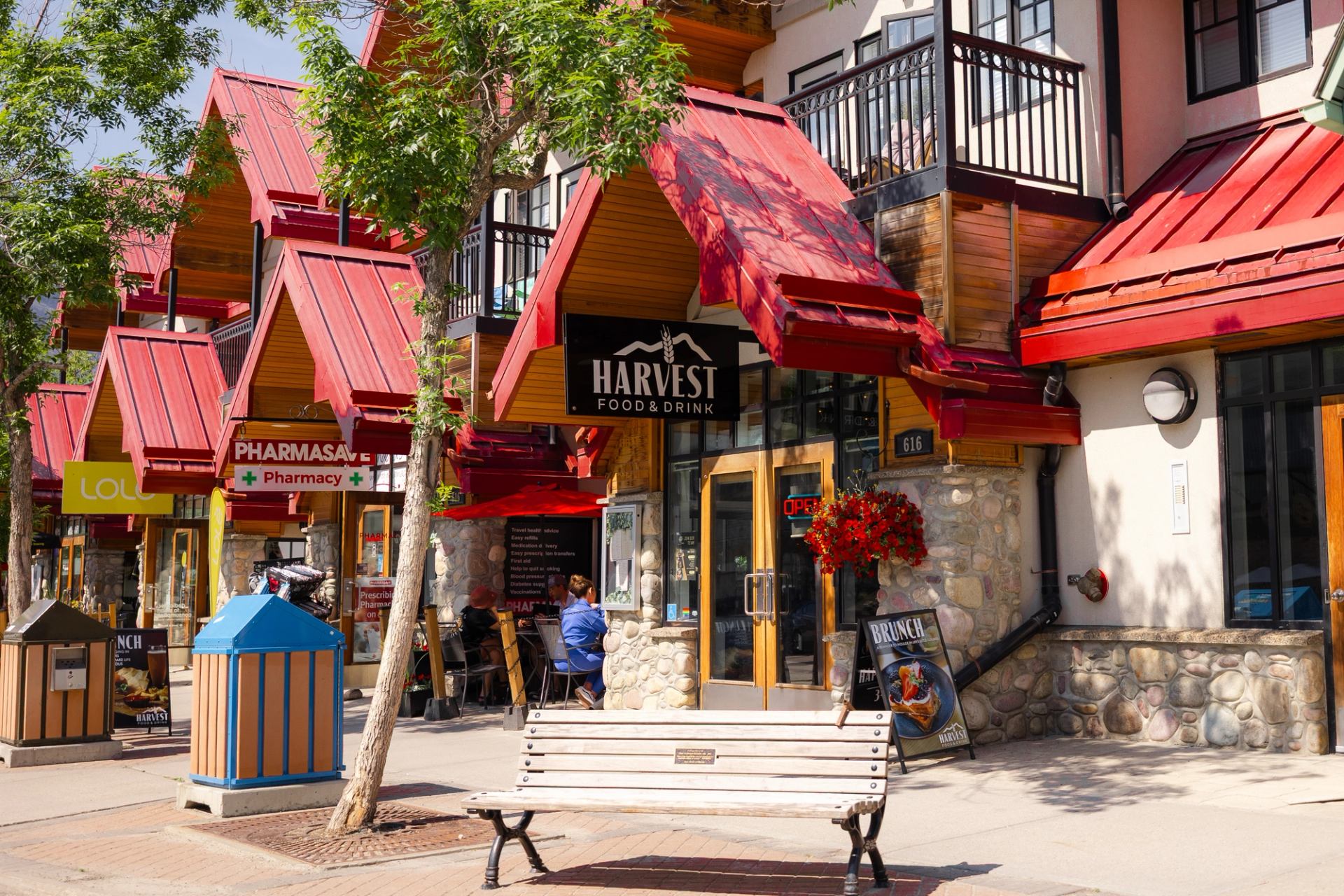 Harvest storefront with red awnings, outdoor seating, and trees along the street.