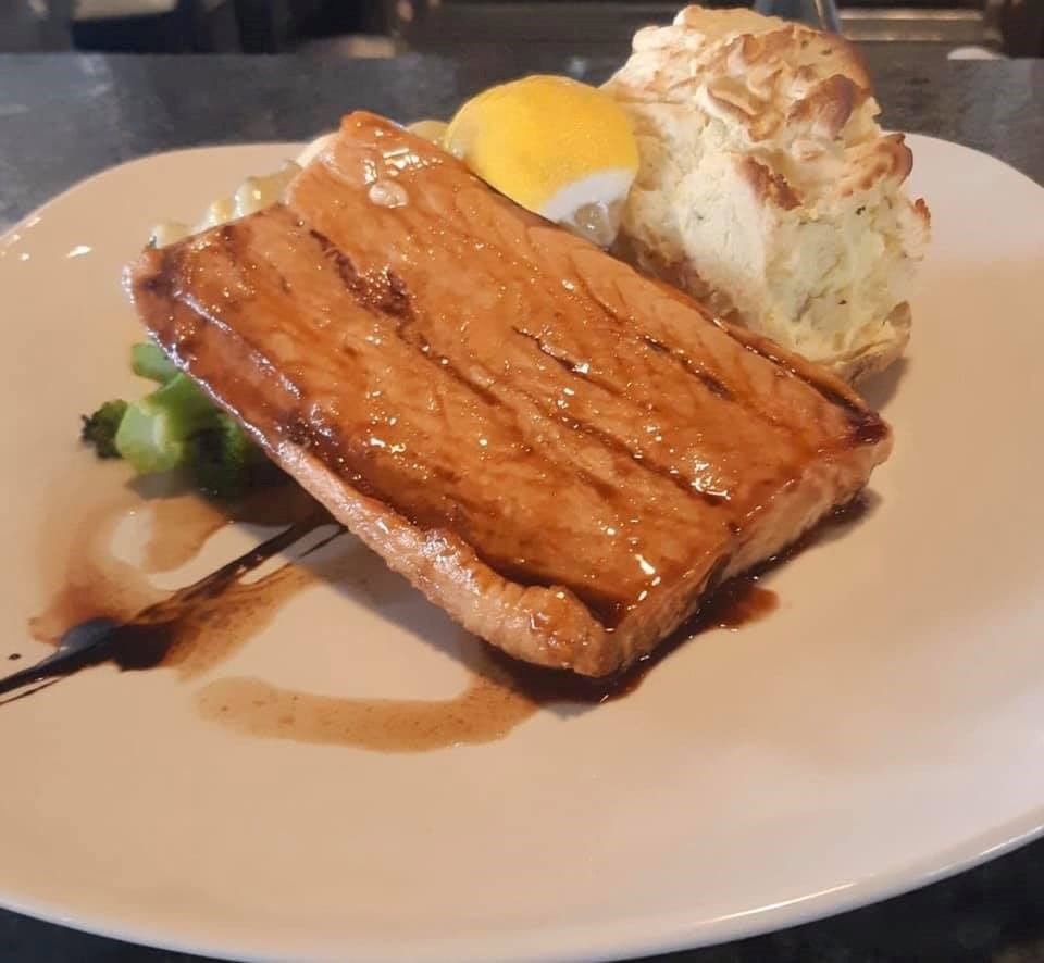 Grilled salmon with greens, lemon wedge, biscuit, and sauce on a white plate.