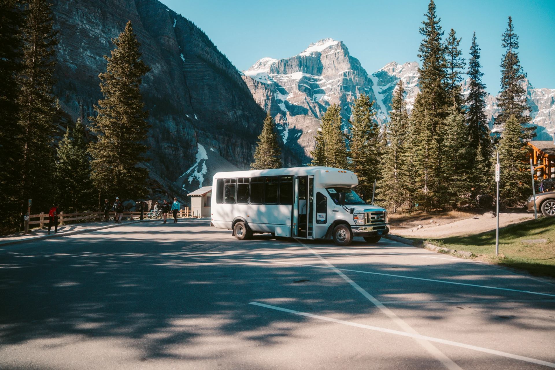 White shuttle bus parked on mountain road with tall evergreen trees and rocky peaks.