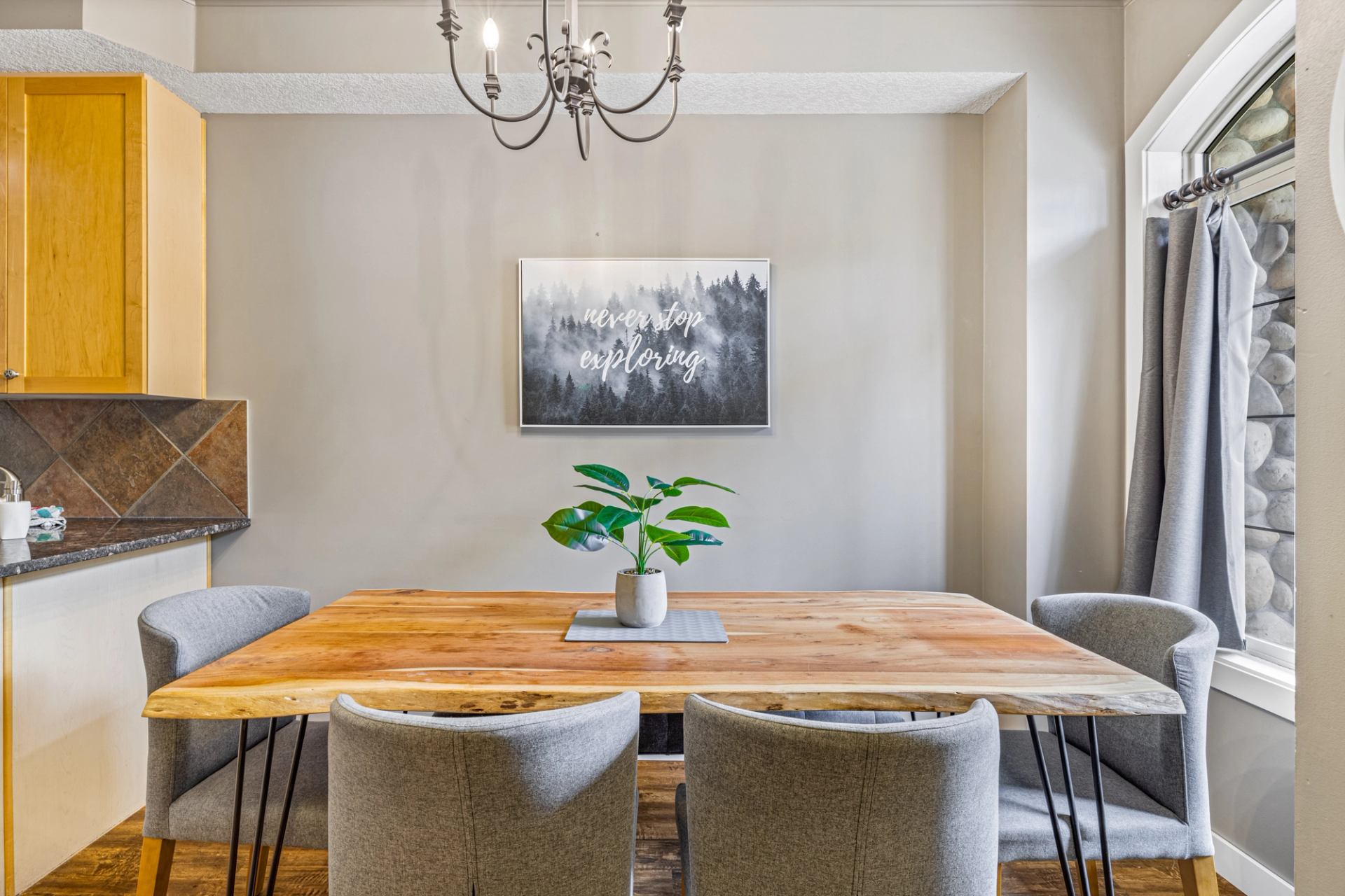 Dining area with live-edge wood table, grey chairs, potted plant and chandelier.
