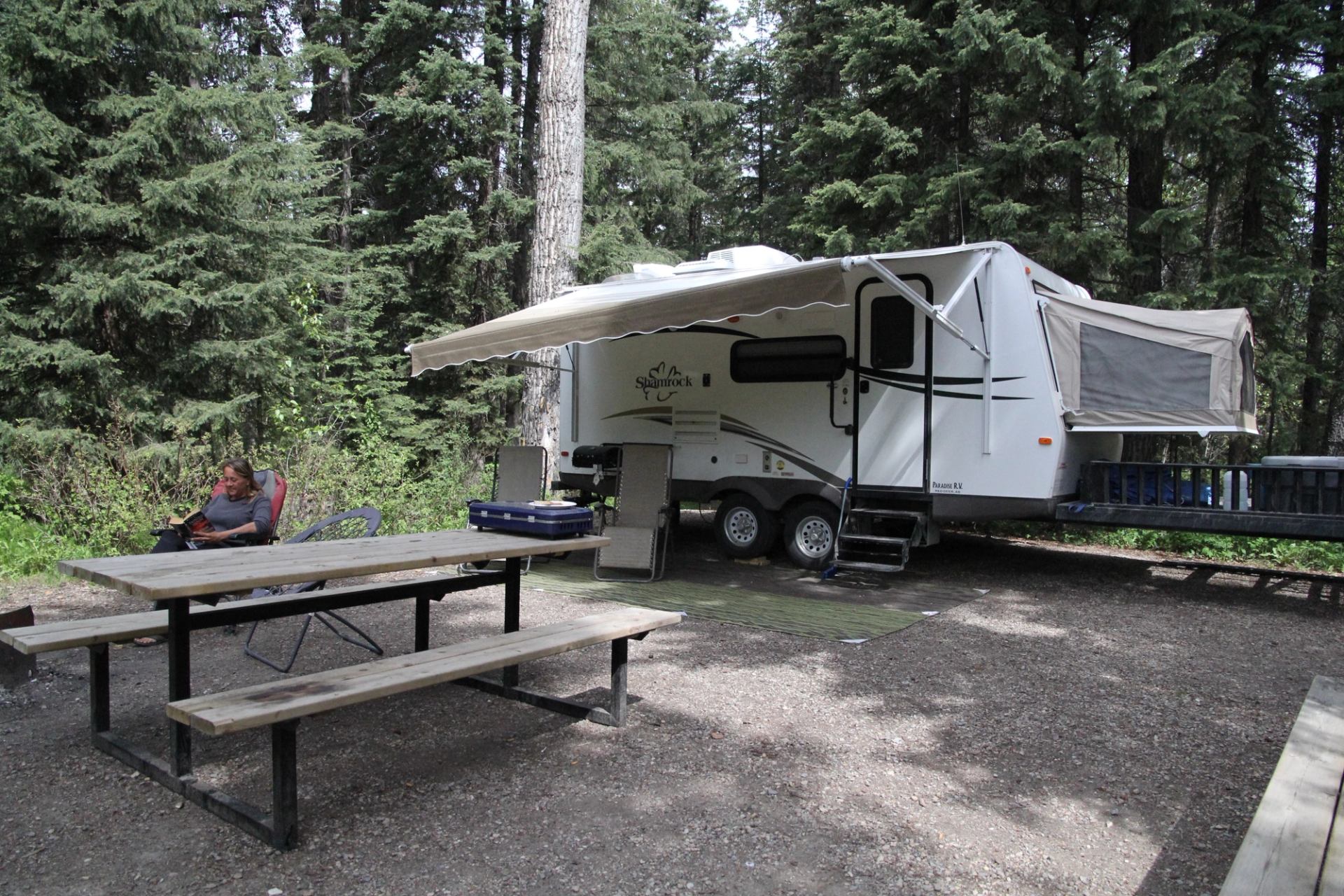 Campsite with travel trailer, extended awning, picnic table and person sitting in chair.