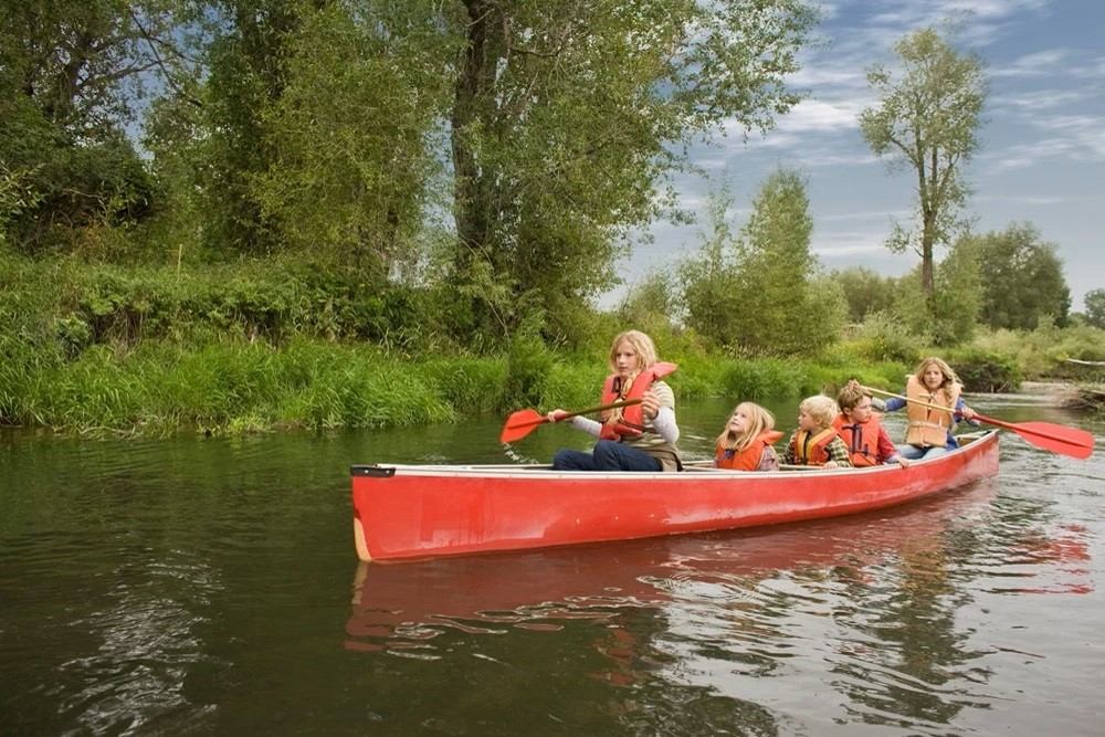 Group paddling red canoe on calm river with green trees and cloudy sky in background.