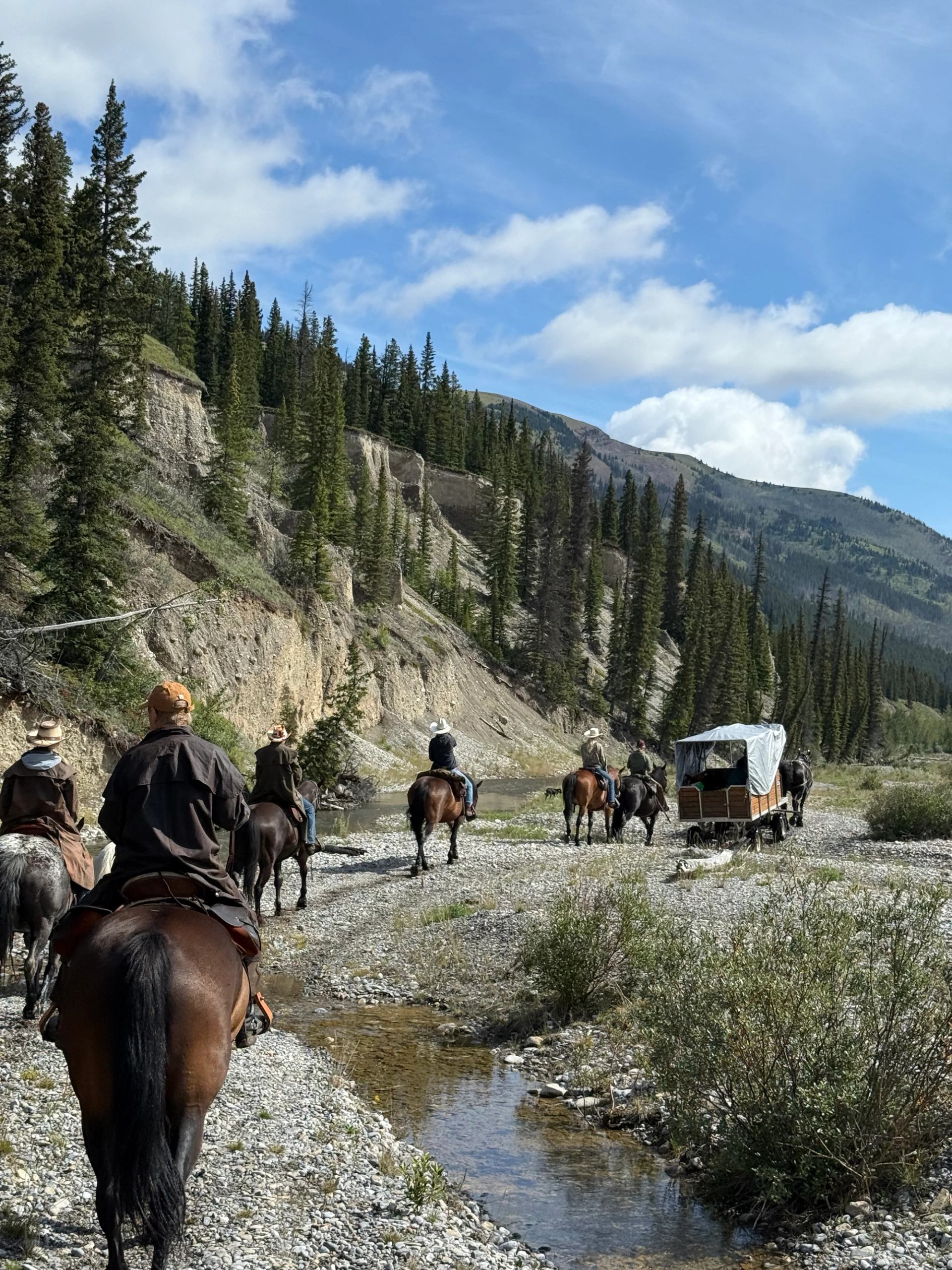 A group of riders and a covered wagon traveling along a rocky mountain valley trail.