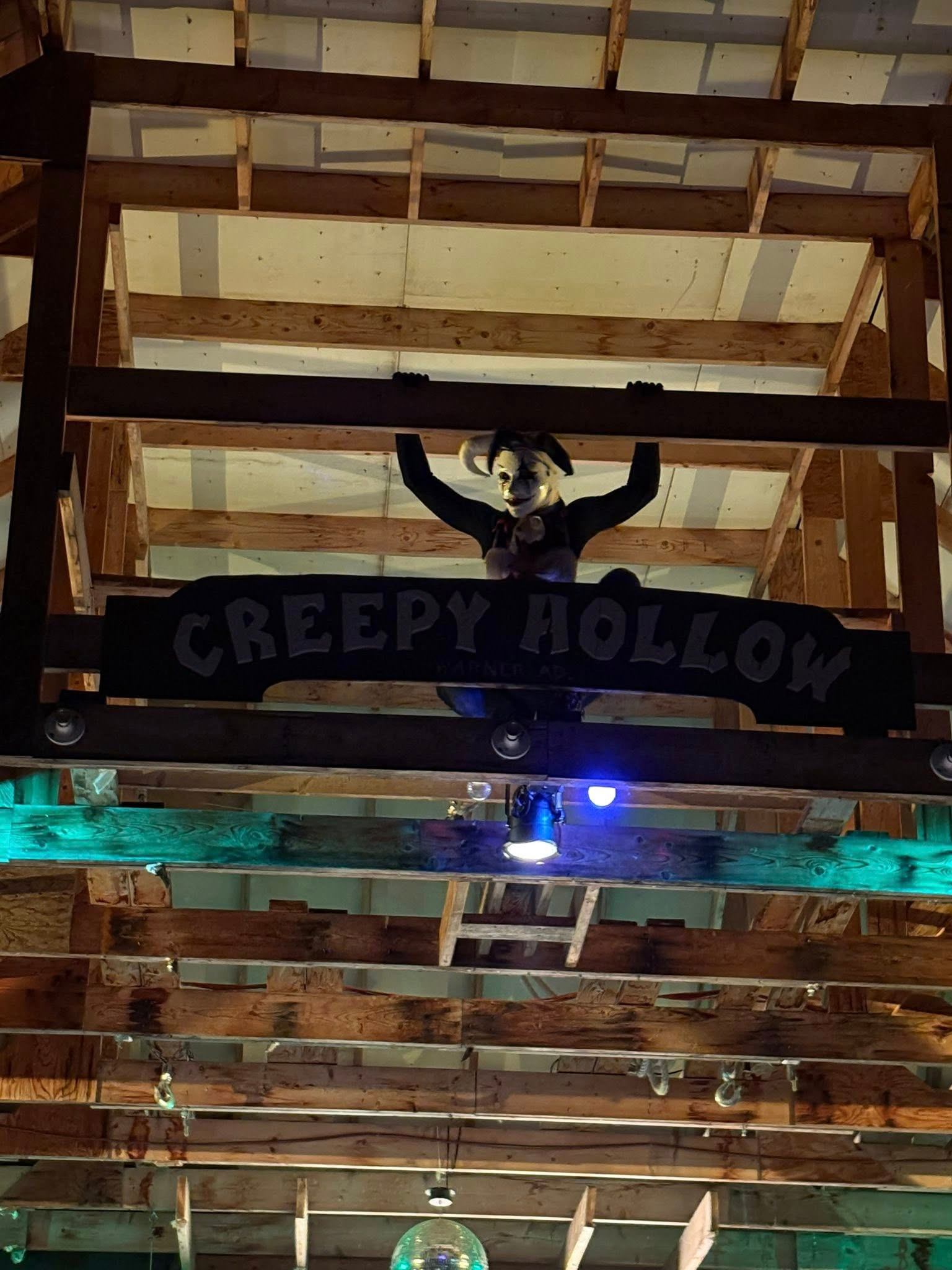 A creepy jester figure hangs from wooden beams above a "CREEPY HOLLOW" sign in a dimly lit, rustic interior.