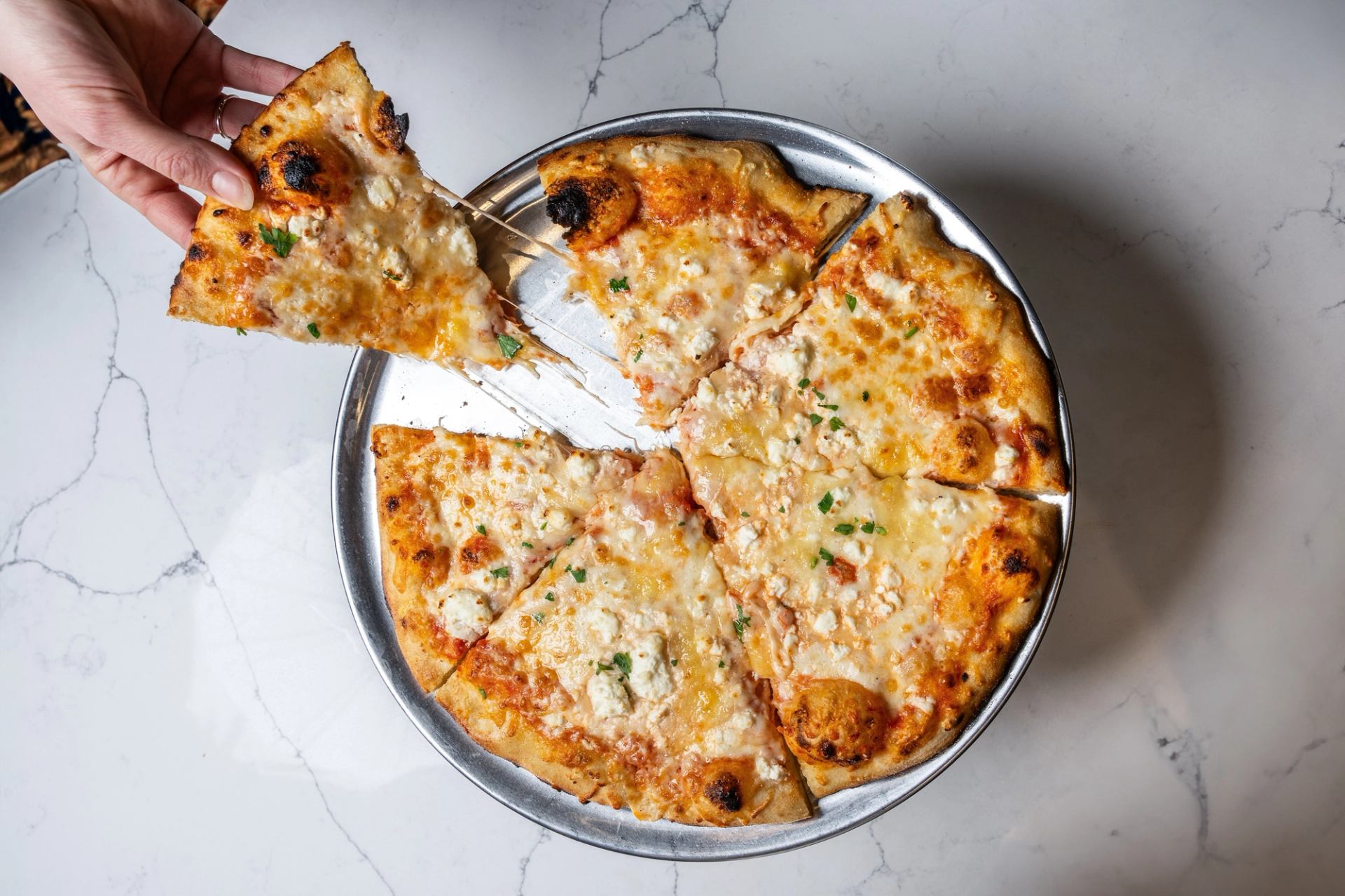 A hand lifting a slice from a baked cheese pizza on a metal tray.