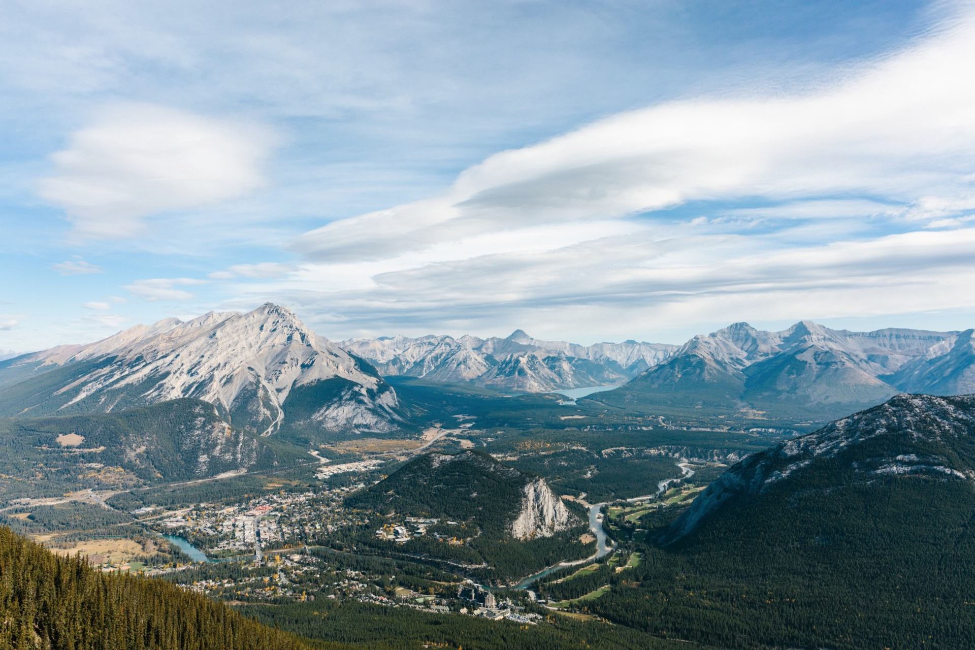 Mountain valley with town, winding river, dense forests and layered peaks under streaked clouds.