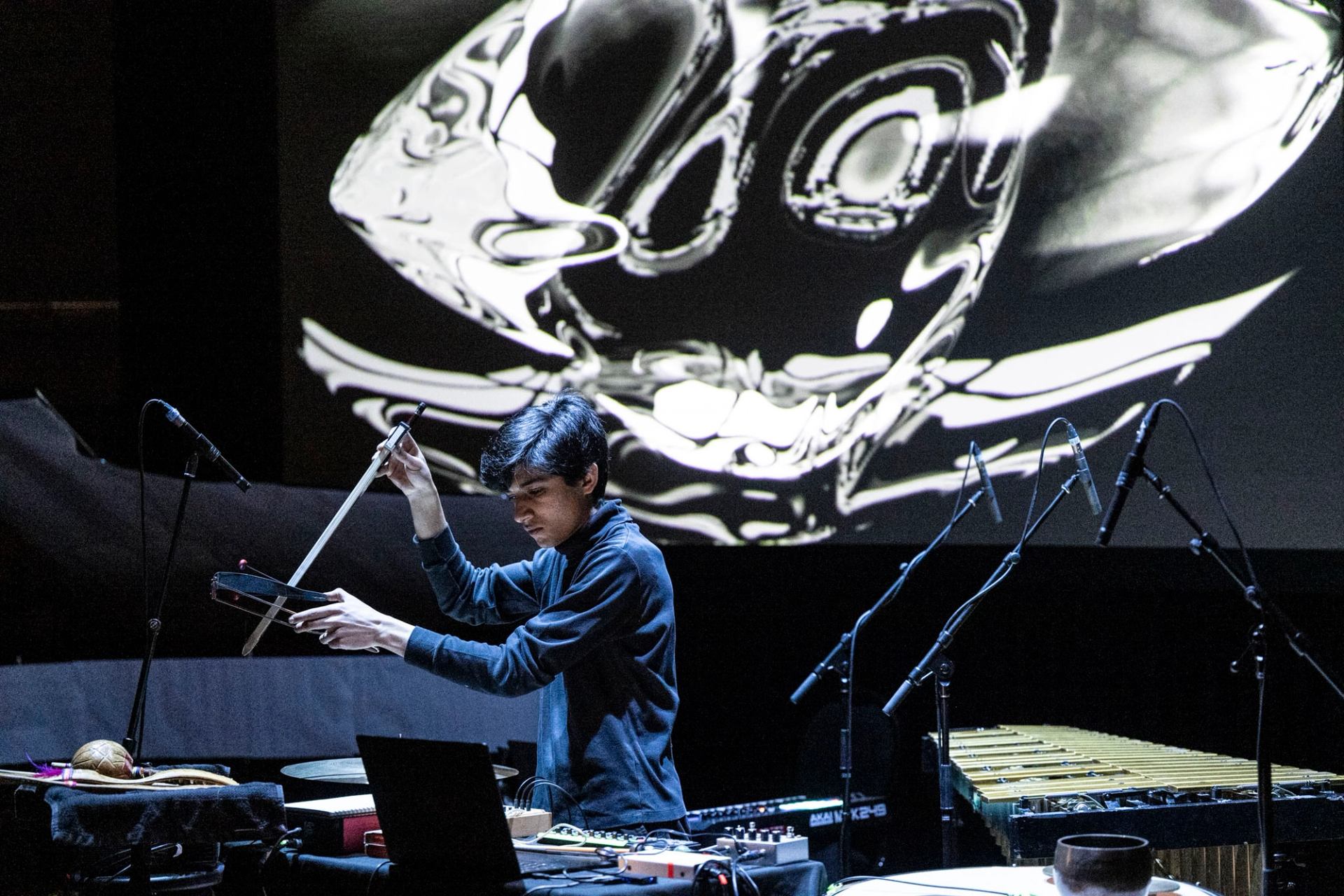 A musician performs with electronic instruments and percussion onstage with abstract visuals projected behind.