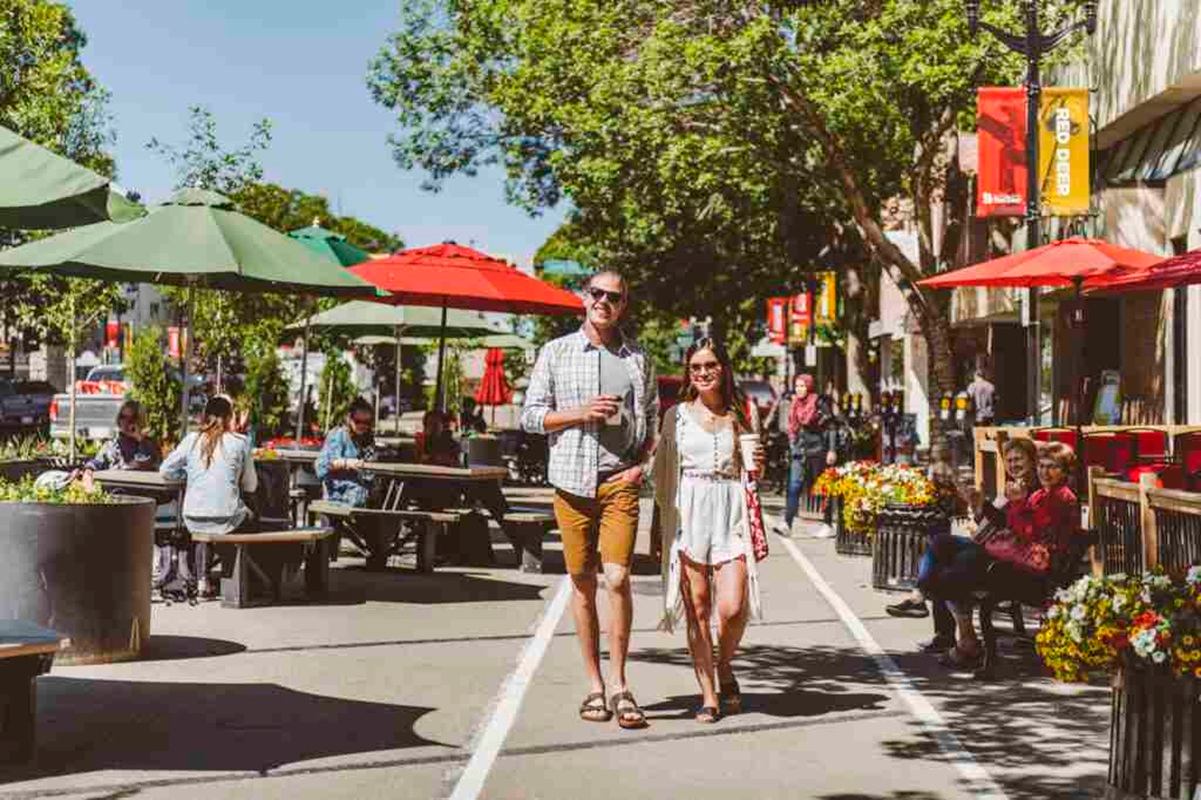 Pedestrians walk past patio tables and umbrellas on a tree-lined city street.