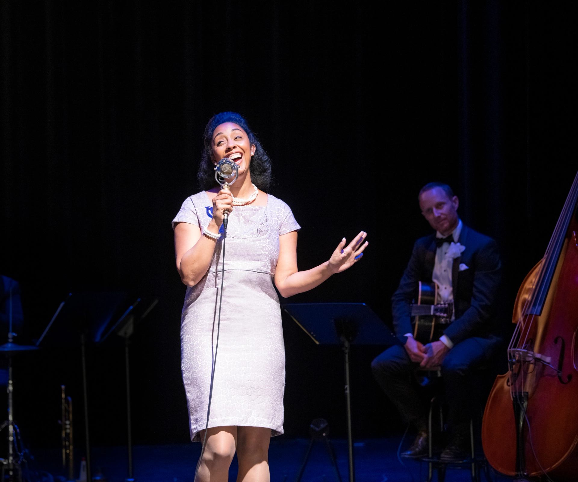 Singer in a light dress performs on stage with guitarist and upright bass nearby.