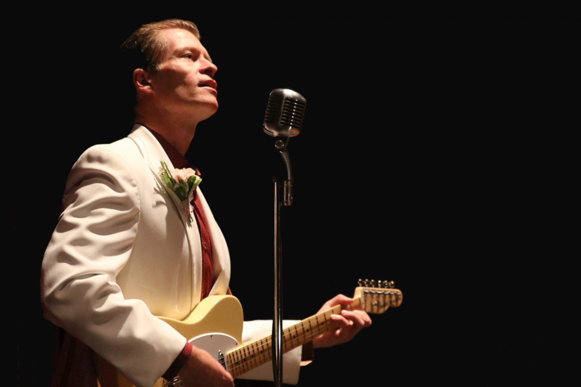 Performer in white suit plays electric guitar beside vintage microphone.