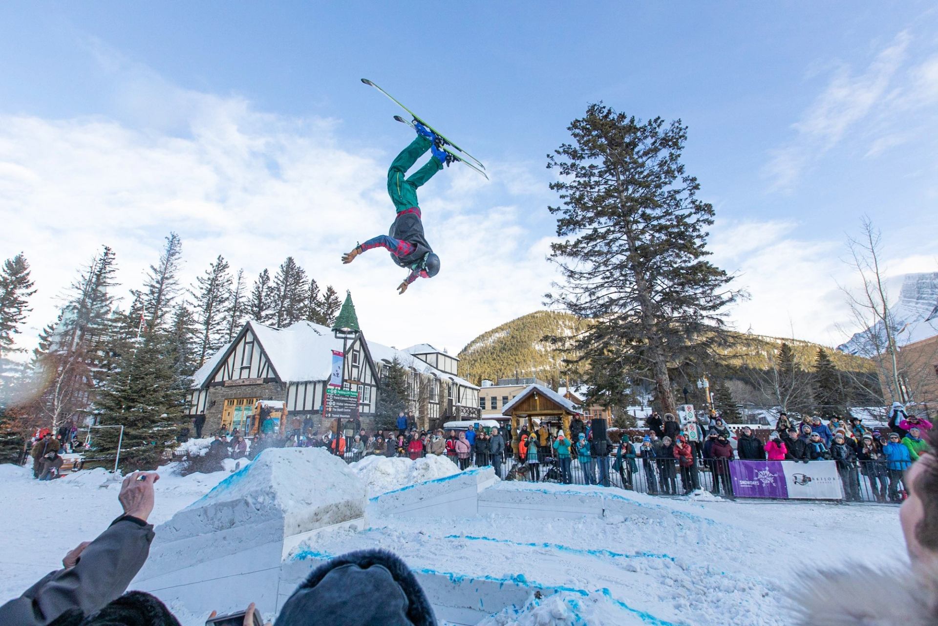 Skier mid-backflip in snowy area with crowd watching.