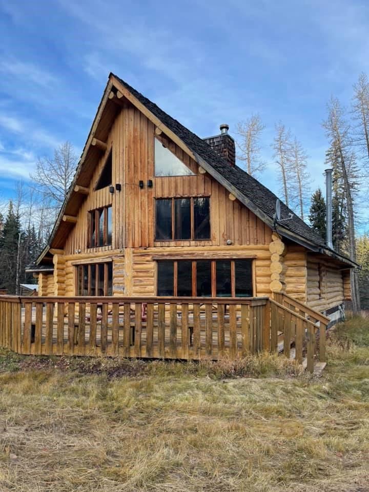 Log cabin with fenced deck and windows, with tall trees in background.