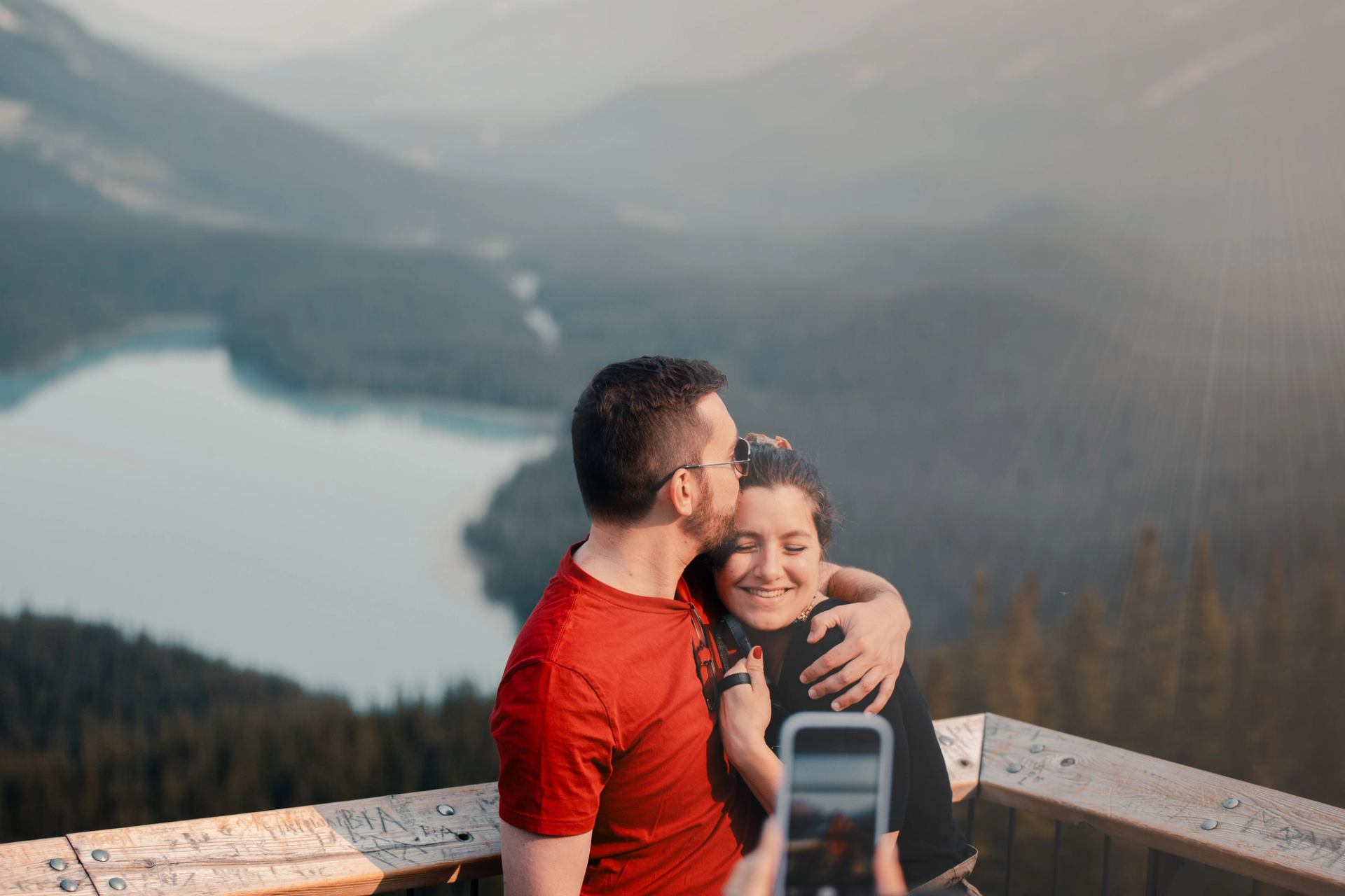 Two people hug on a wooden lookout above a lake, with phone taking photo.