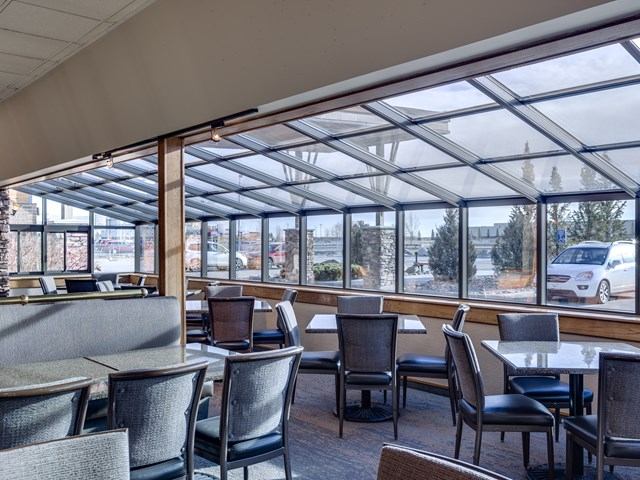 Hotel dining area with tables and chairs beside large glass windows, overlooking a parking area and trees.