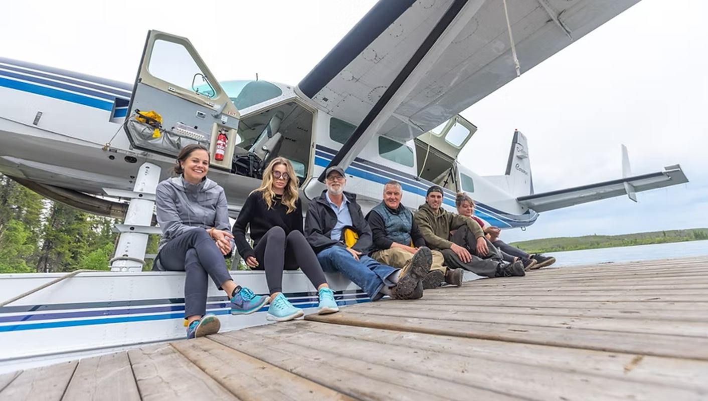 Group of people sit on dock beside floatplane, with lake and trees in background.