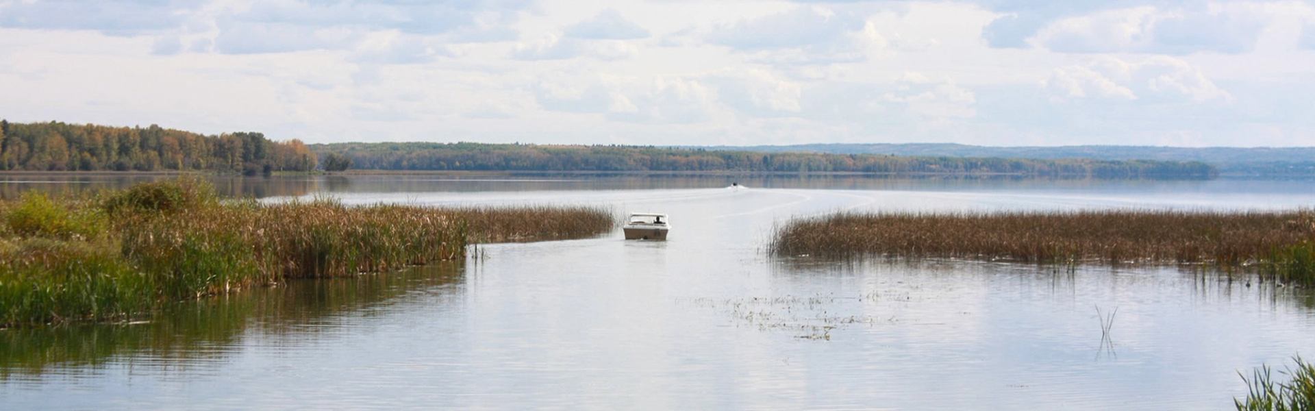 A boat floats near reeds on Buck Lake with forested hills in the distance under a cloudy sky.