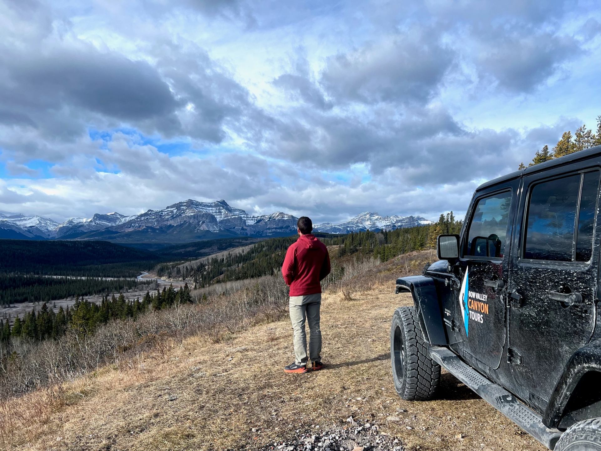 Person stands on a ridge overlooking mountains with a black Jeep parked nearby.