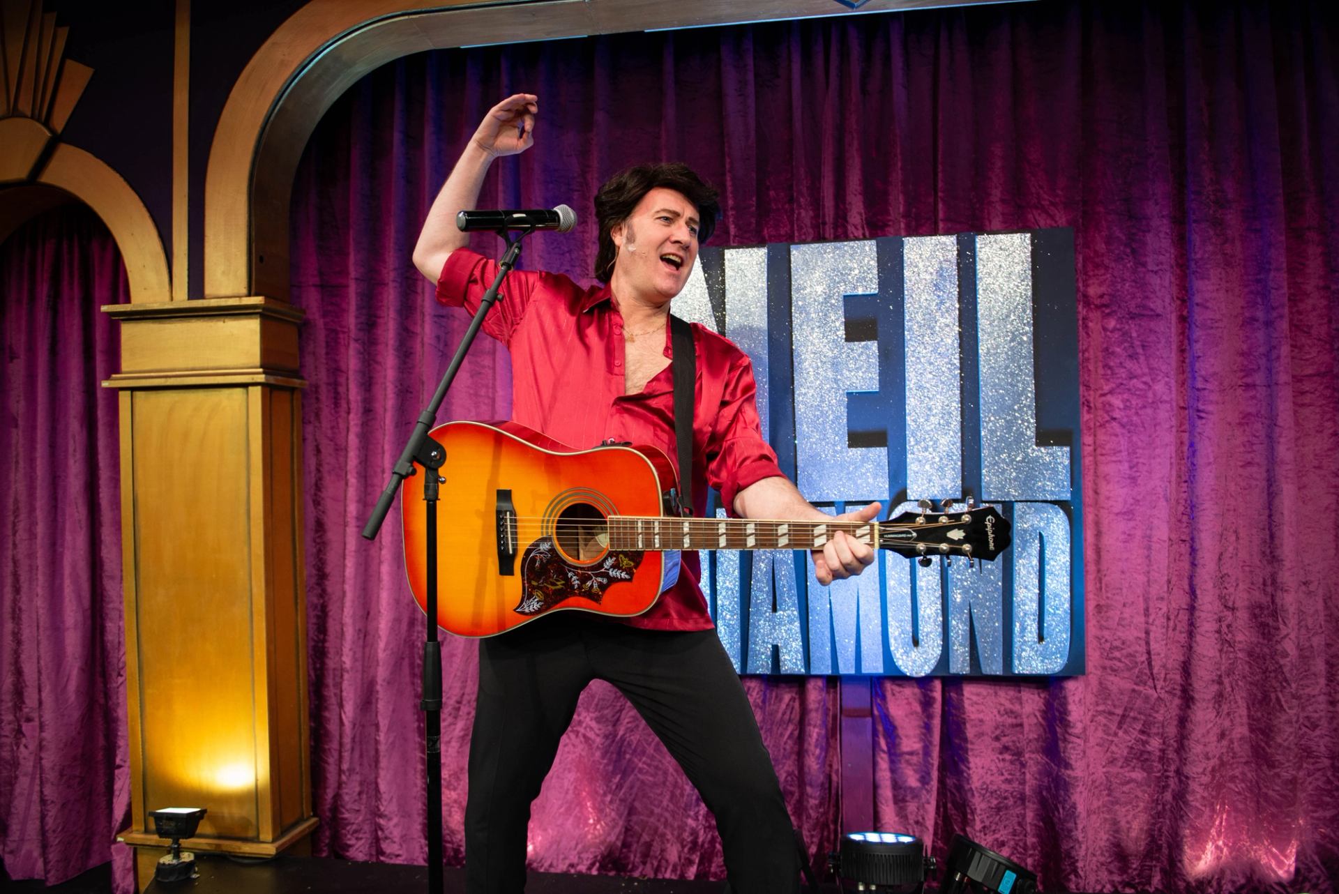 Guitarist in red shirt performs on stage before glittery Neil Diamond backdrop and curtains.