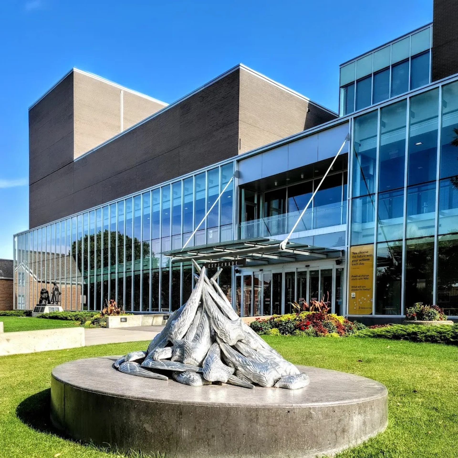 Metal bird sculpture on circular pedestal outside modern glass building with landscaped lawn.