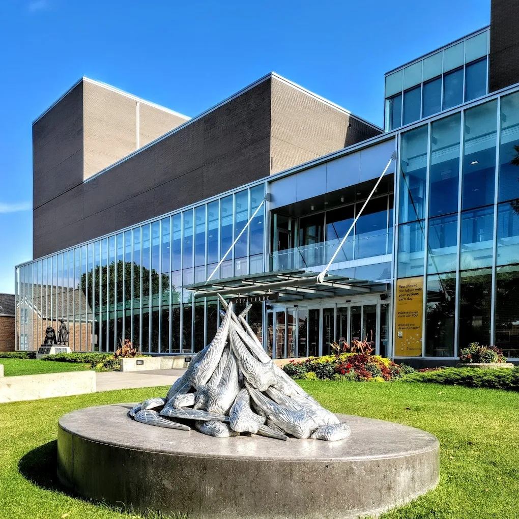 Metal bird sculpture on circular pedestal outside modern glass building with landscaped lawn.