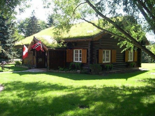 Traditional log house with grass roof, flags, and orange shutters in green setting.