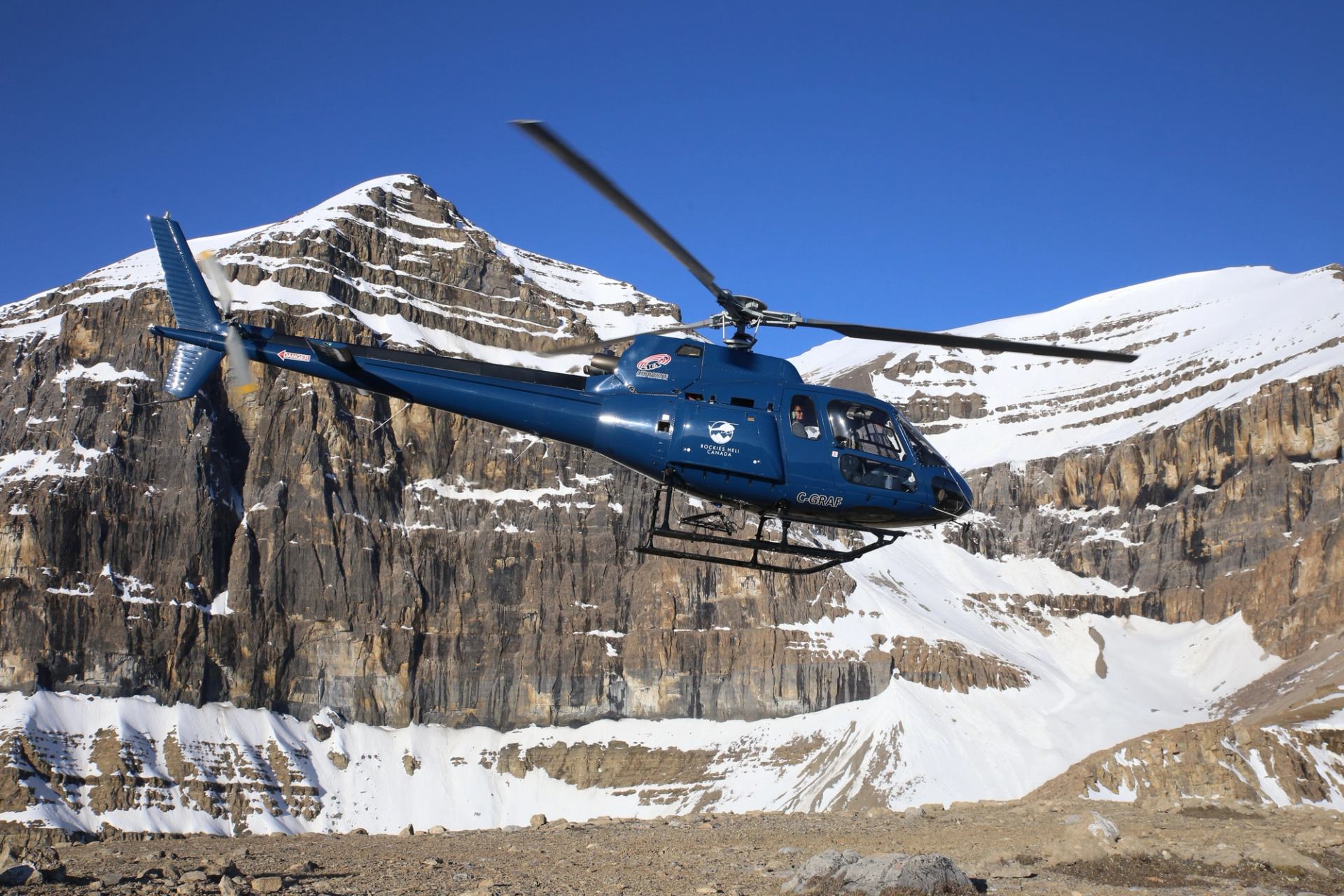 Blue helicopter flying past rugged snow covered mountain peaks.