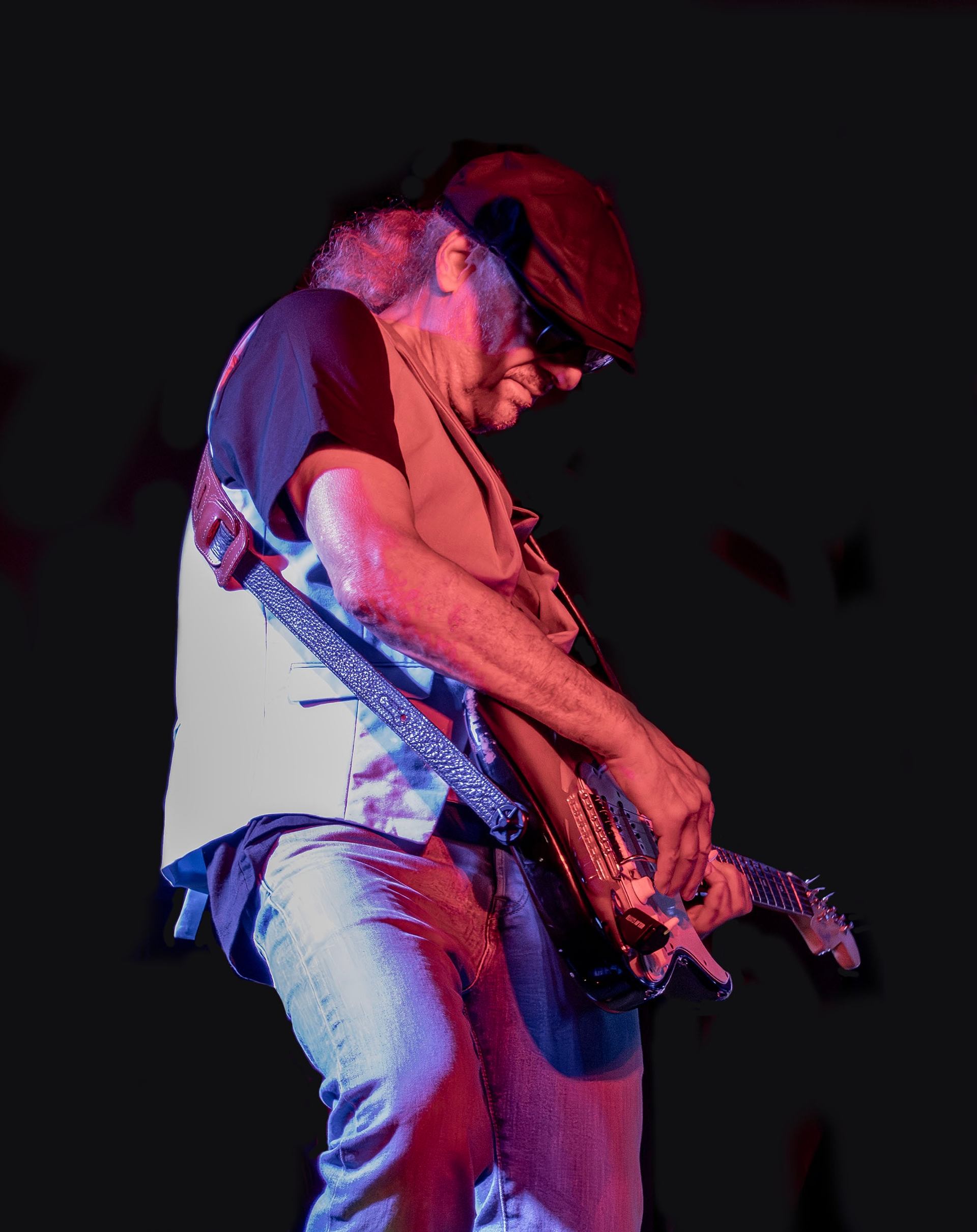 Guitarist performing on stage under red and blue lighting, wearing a cap and vest while playing an electric guitar.