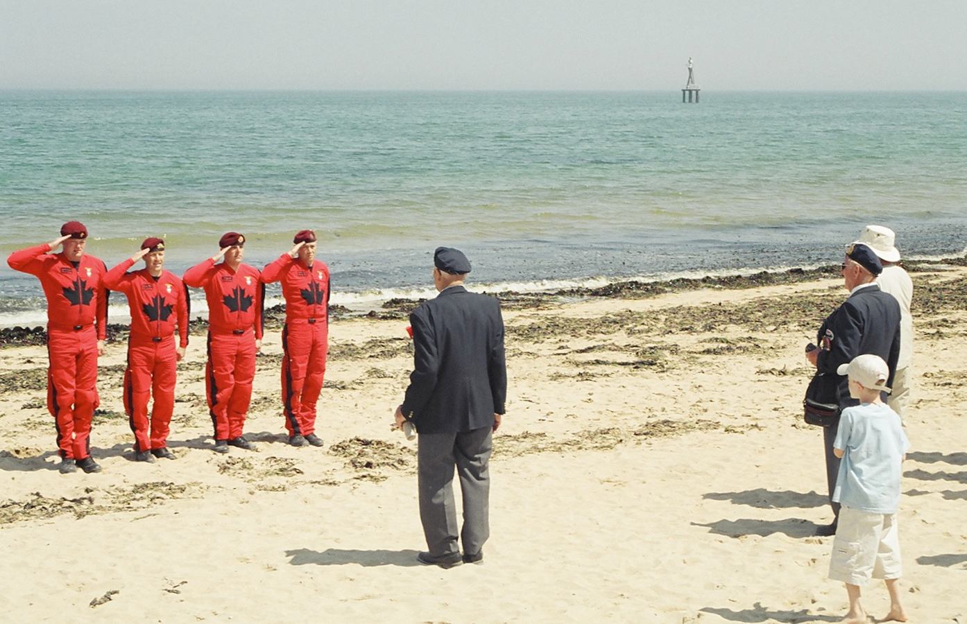 Canadian paratroopers in red uniforms salute veterans and children on a sandy beach.