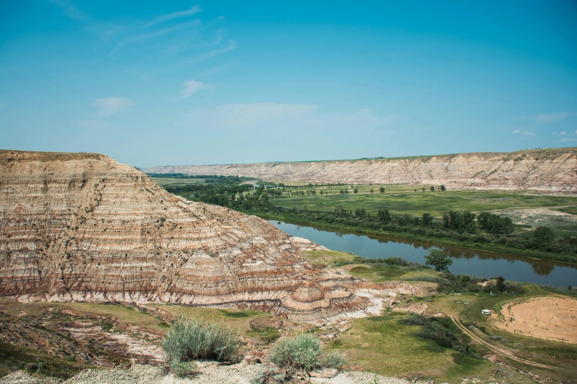 Striped badlands cliffs overlook a winding river and green valley under clear sky.