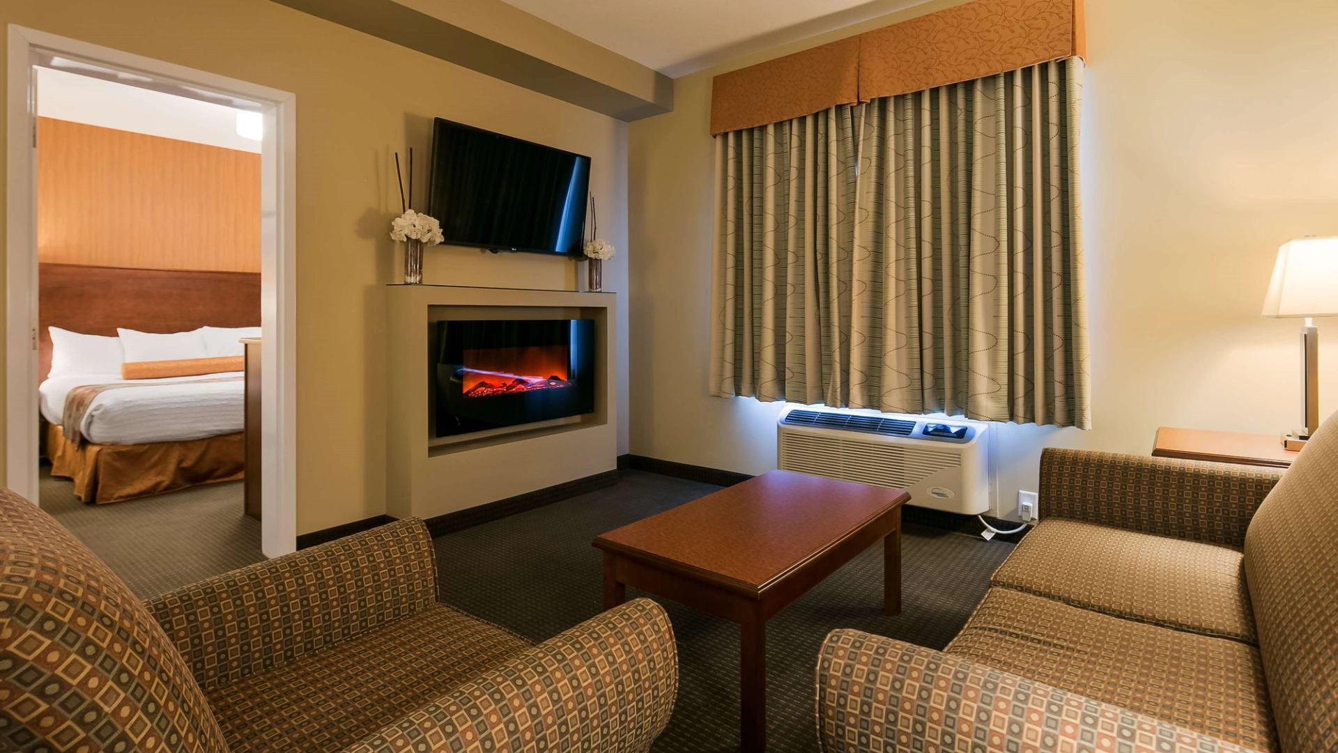 Hotel suite living area with sofa, armchair, TV, fireplace, and doorway to bedroom.