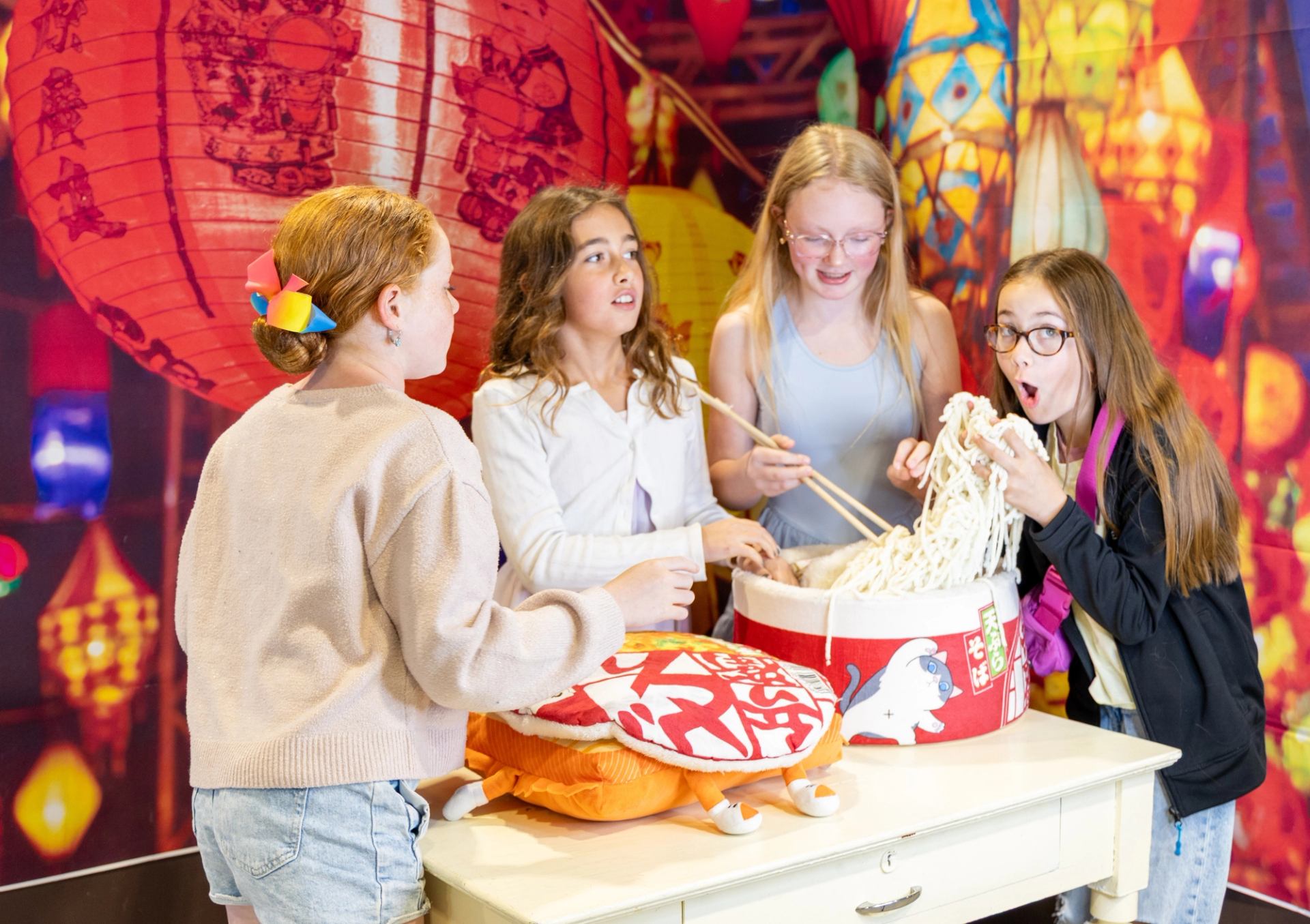 Children play with oversized noodle props in front of colourful lantern backdrop.