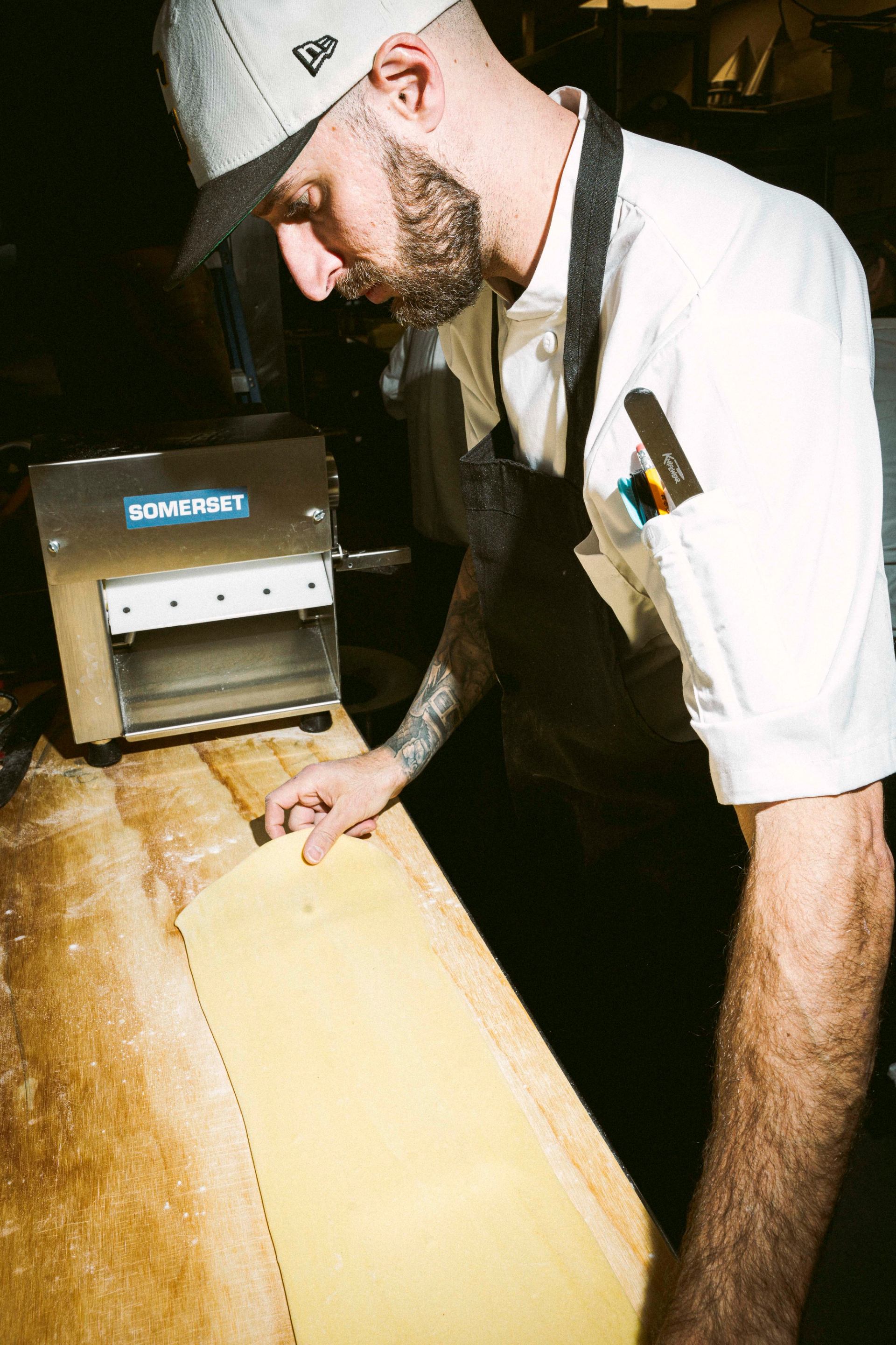A bearded chef in a cap works with a sheet of pasta dough on a floured wooden table next to a pasta machine.