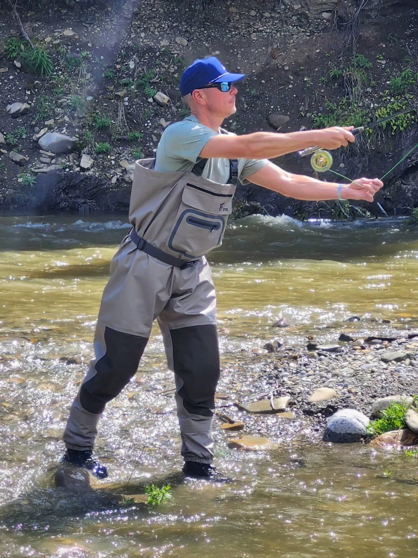 Person in waders fly fishes in shallow rocky river near steep, vegetated bank.
