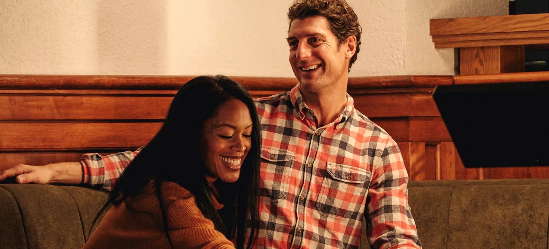 Two people sit on a couch, talking together in a wood-panelled room.