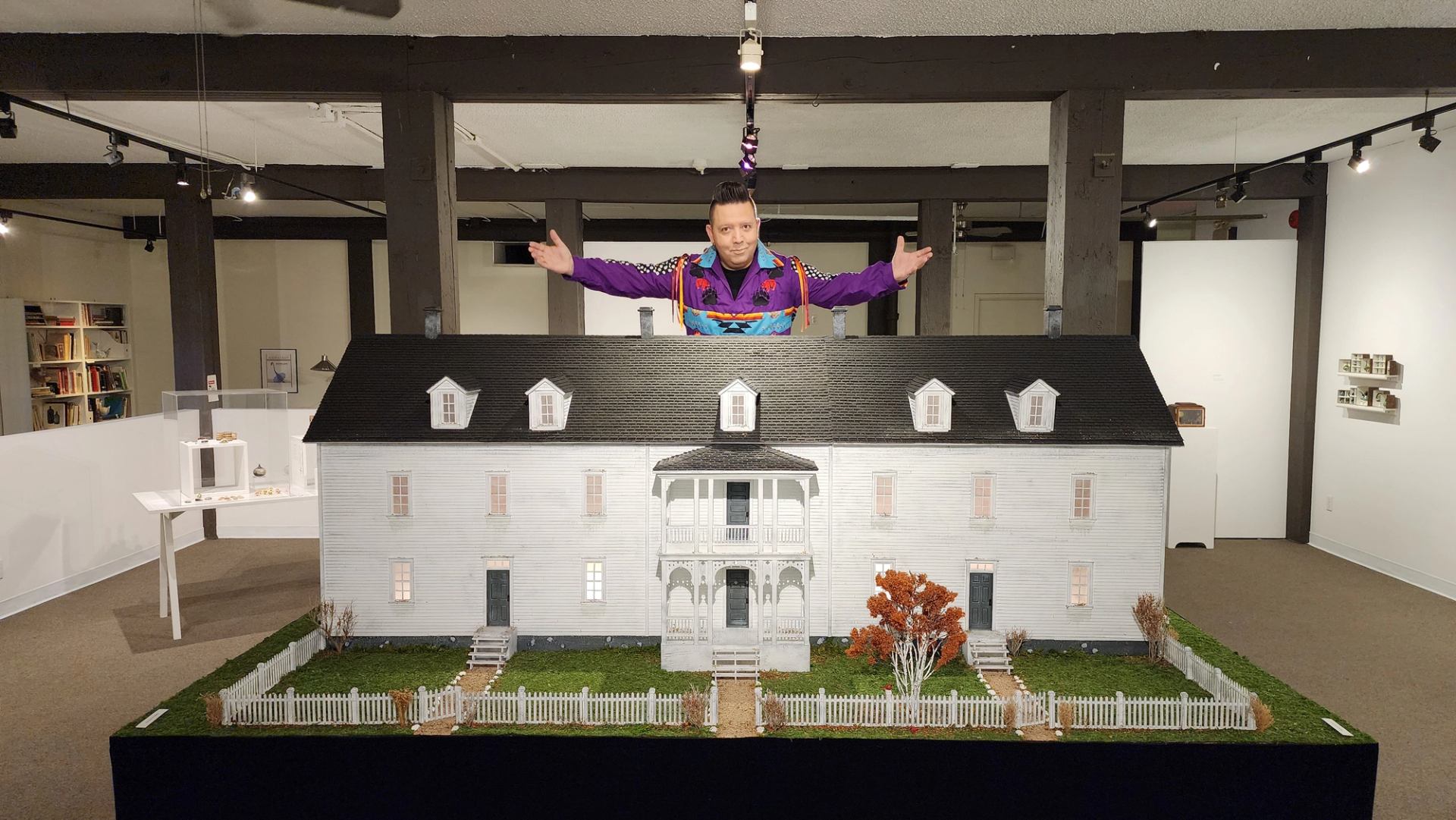 Man in colourful ceremonial outfit with arms out, standing behind miniature house exhibit indoors.