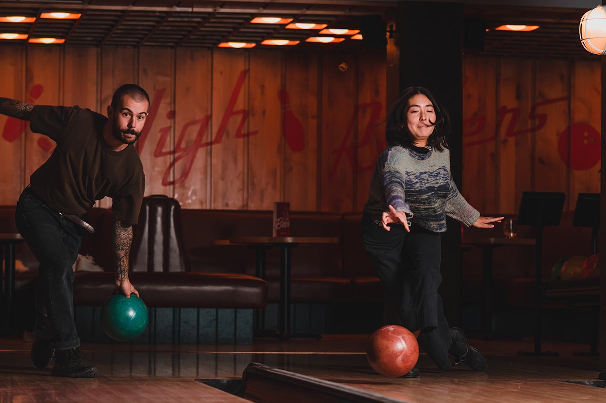 A smiling woman releases a red bowling ball as a man prepares to bowl with a green ball in a dimly lit alley.