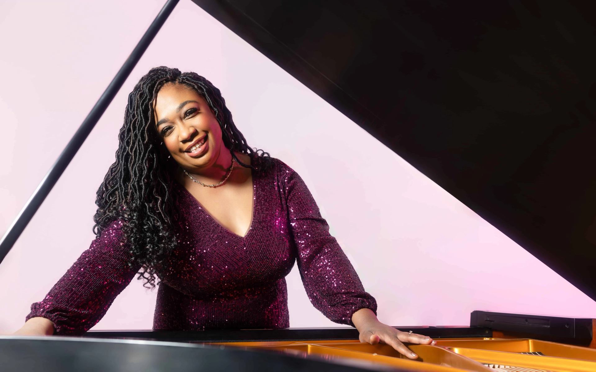 Person in sequined purple top leaning over grand piano with pink background.