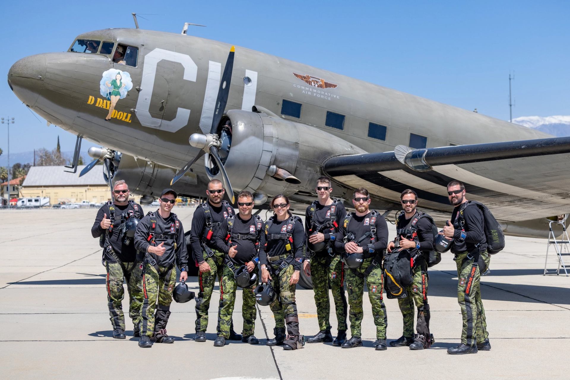 Parachute team in camouflage stands in front of vintage military aircraft on tarmac.