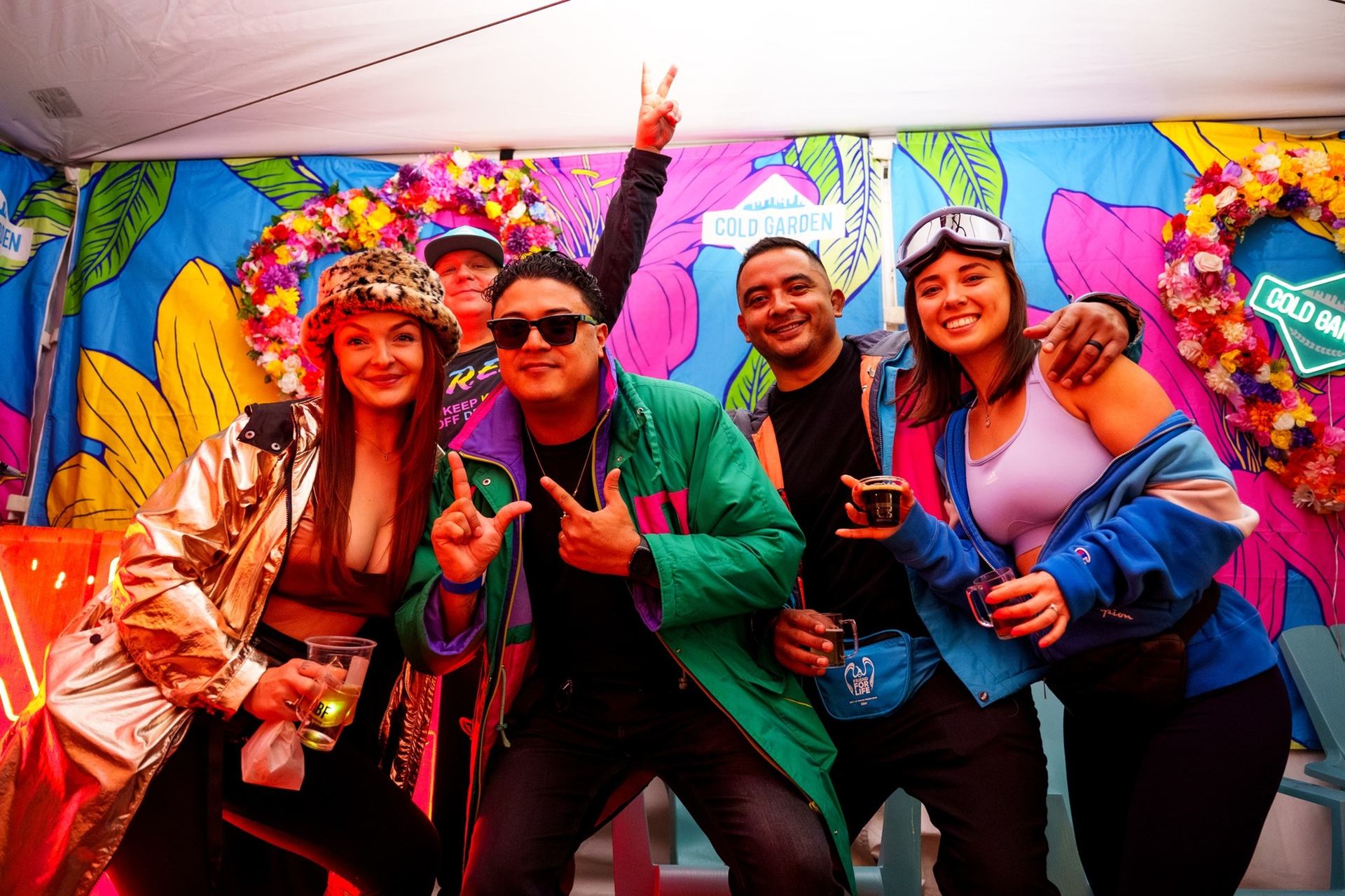 Group of friends posing in colourful jackets before Cold Garden floral backdrop at event.