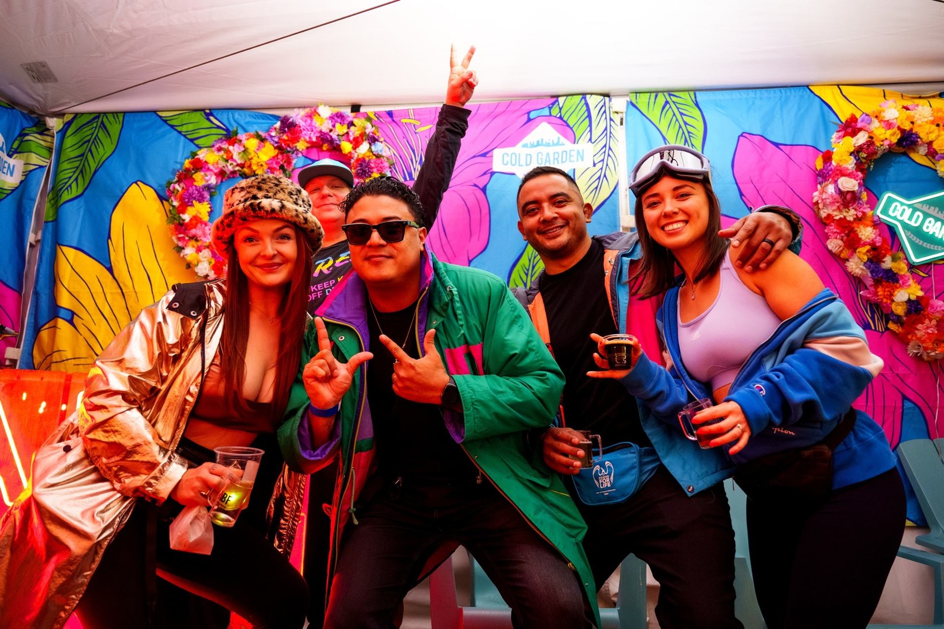Group of friends posing in colourful jackets before Cold Garden floral backdrop at event.
