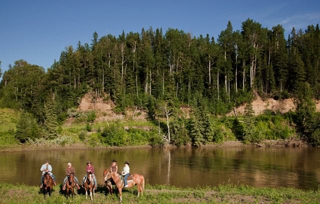Riverwood Ranch Trail Ride, riders on horseback beside a calm river and forested hillside.