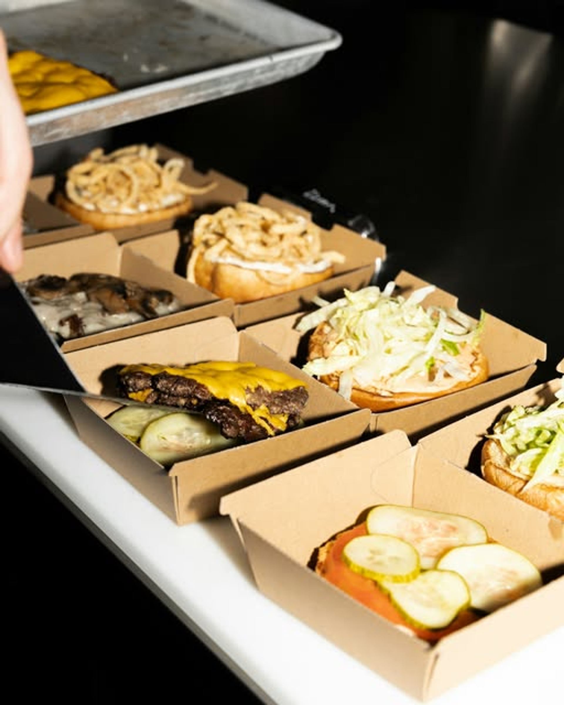 Open burger boxes display cheeseburgers and toppings being assembled on a restaurant prep counter.