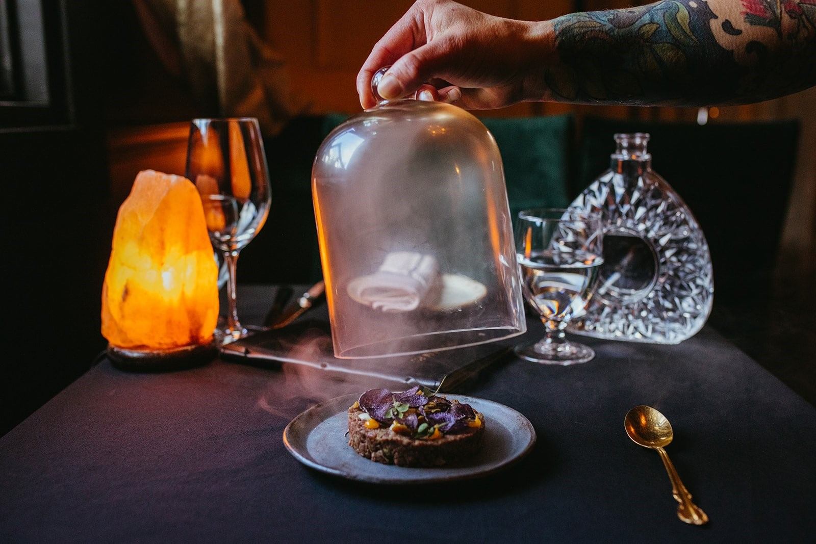A hand lifting a glass cloche over a smoky dish, with elegant glassware and a salt lamp nearby.