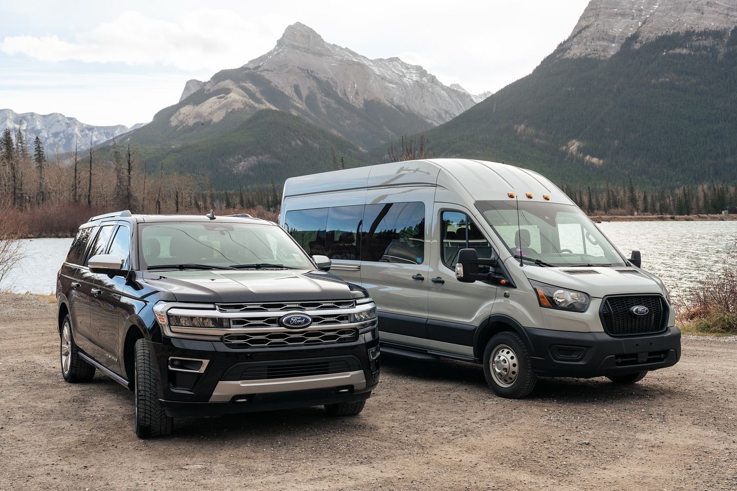 SUV and shuttle van parked by a lake with Rocky Mountains in the background.