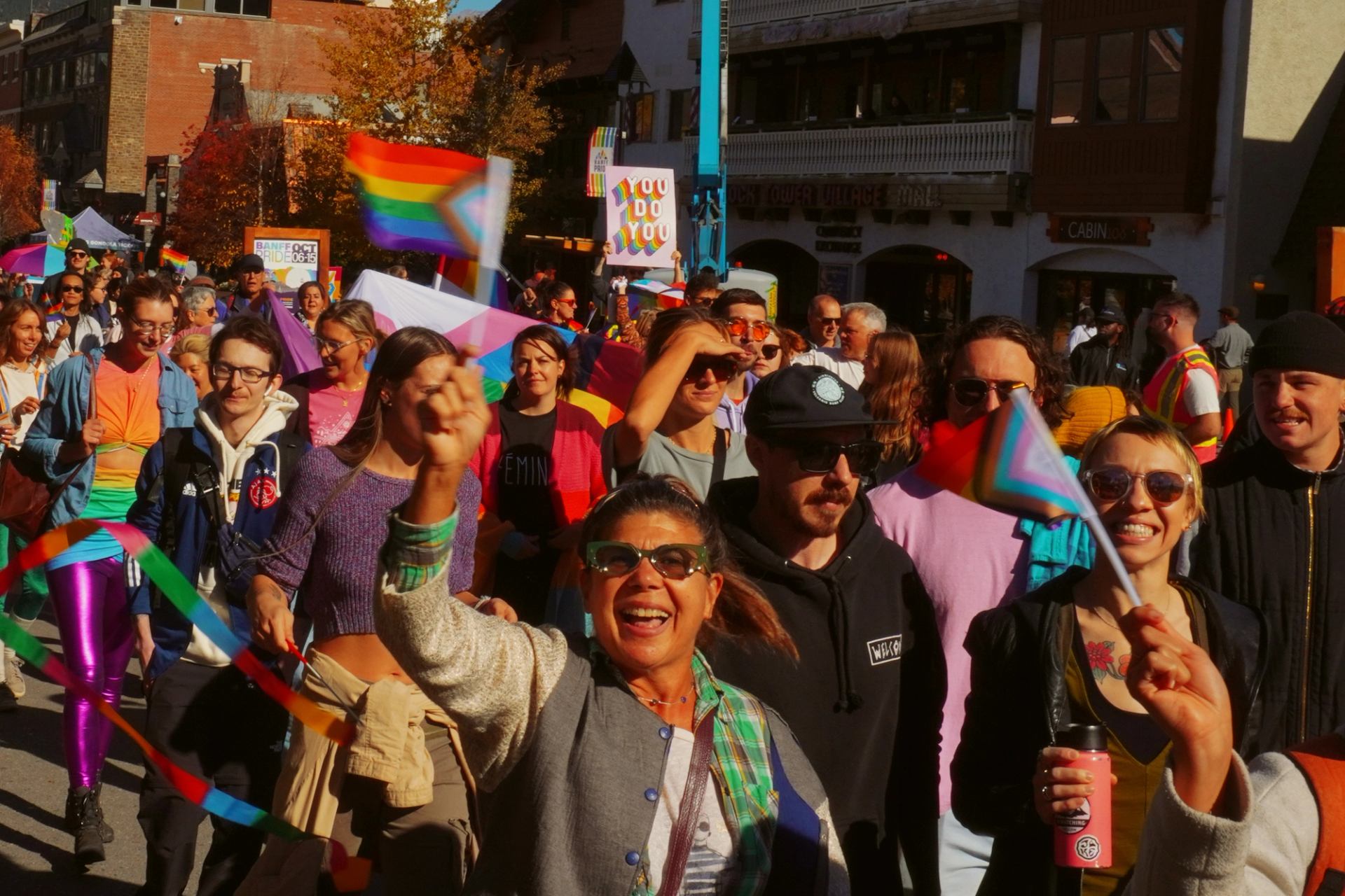 Crowd marches with Pride flags, colourful ribbons and signs along a city street.