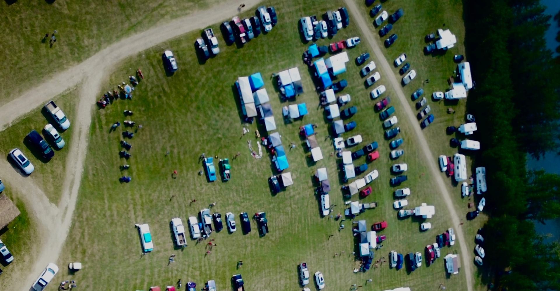 Aerial view of outdoor event with tents, parked vehicles and people on grassy field.