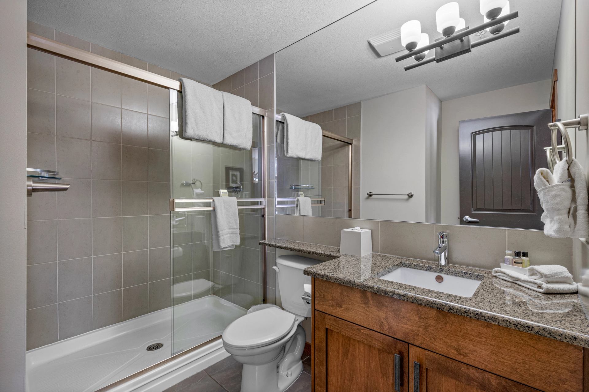 Bathroom with glass shower, toilet, granite vanity, large mirror and white towels.